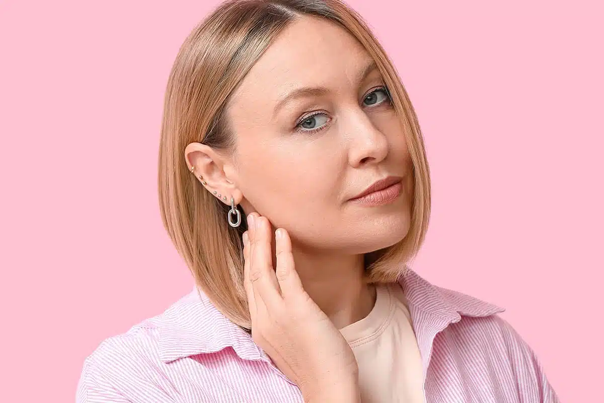 A woman with blonde hair wearing several silver earrings, looking forward while resting her hand near her jawline against a pink background.