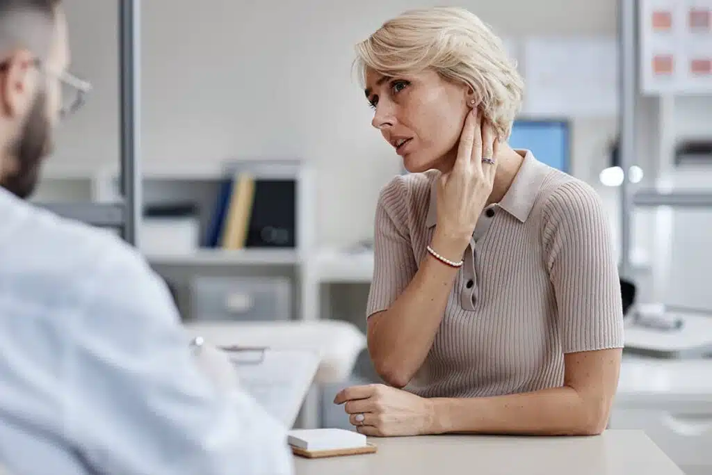 A woman sitting in a medical office, touching the area behind her ear while speaking with a healthcare professional.